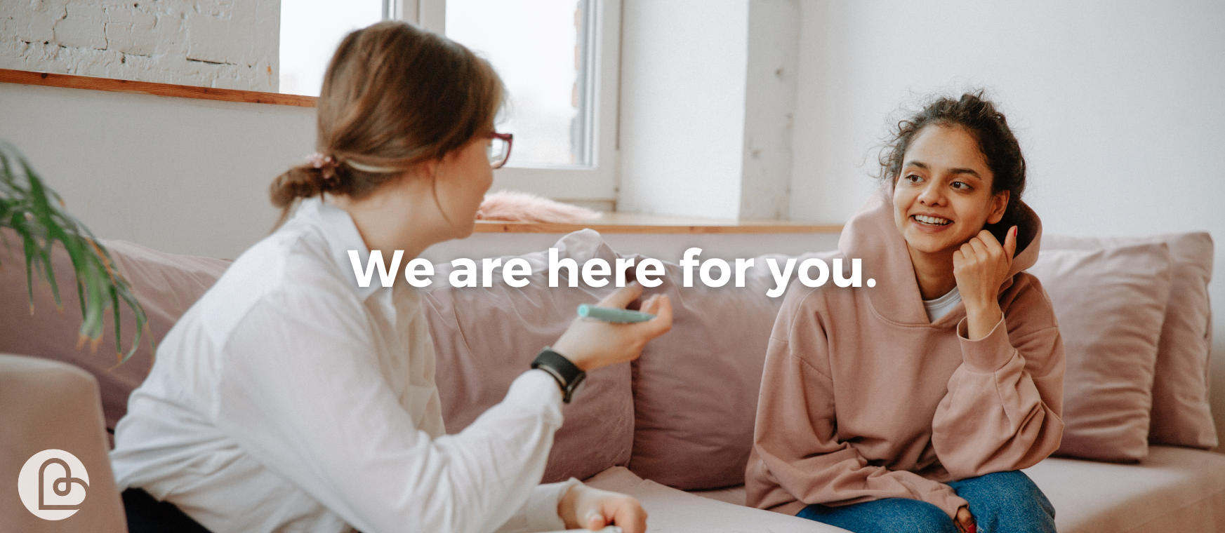Image of a support worker and client sitting on a couch talking. Overlying text reads: We are here for you.